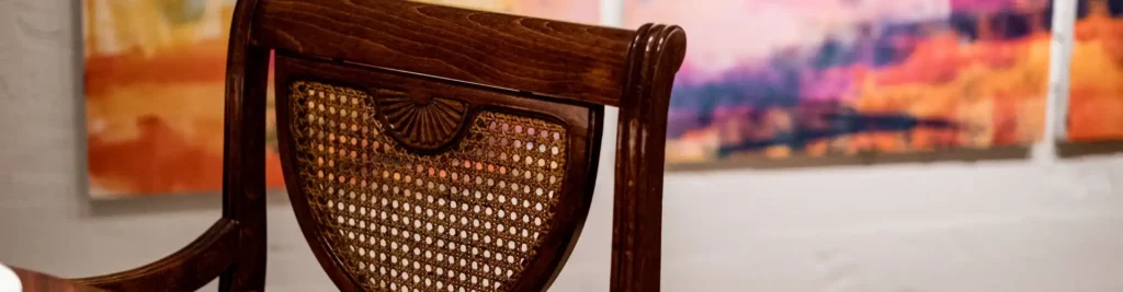 Muestra en primer plano una silla de madera antigua y de fondo desenfocado una pared con un cuadro colorido, para mostrar El espacio del Restaurante Pabu