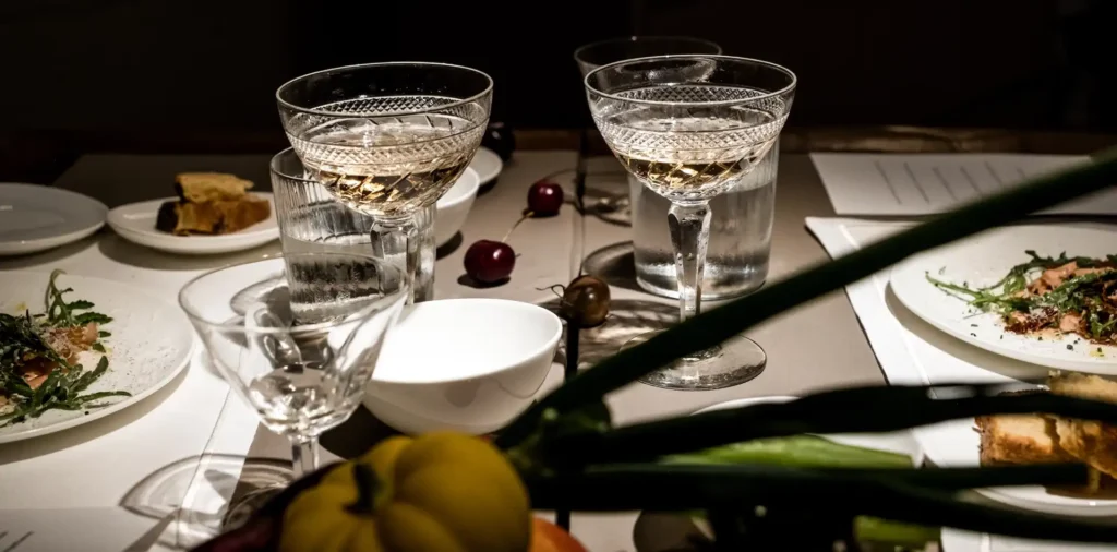 Se ve una mesa con su mantel con dos copas con vino blanco y varios platos de comida