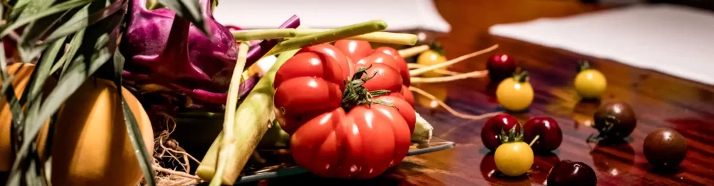 La foto muestra diferentes verduras encima de una mesa de madera y en el centro un tomate rojo grande